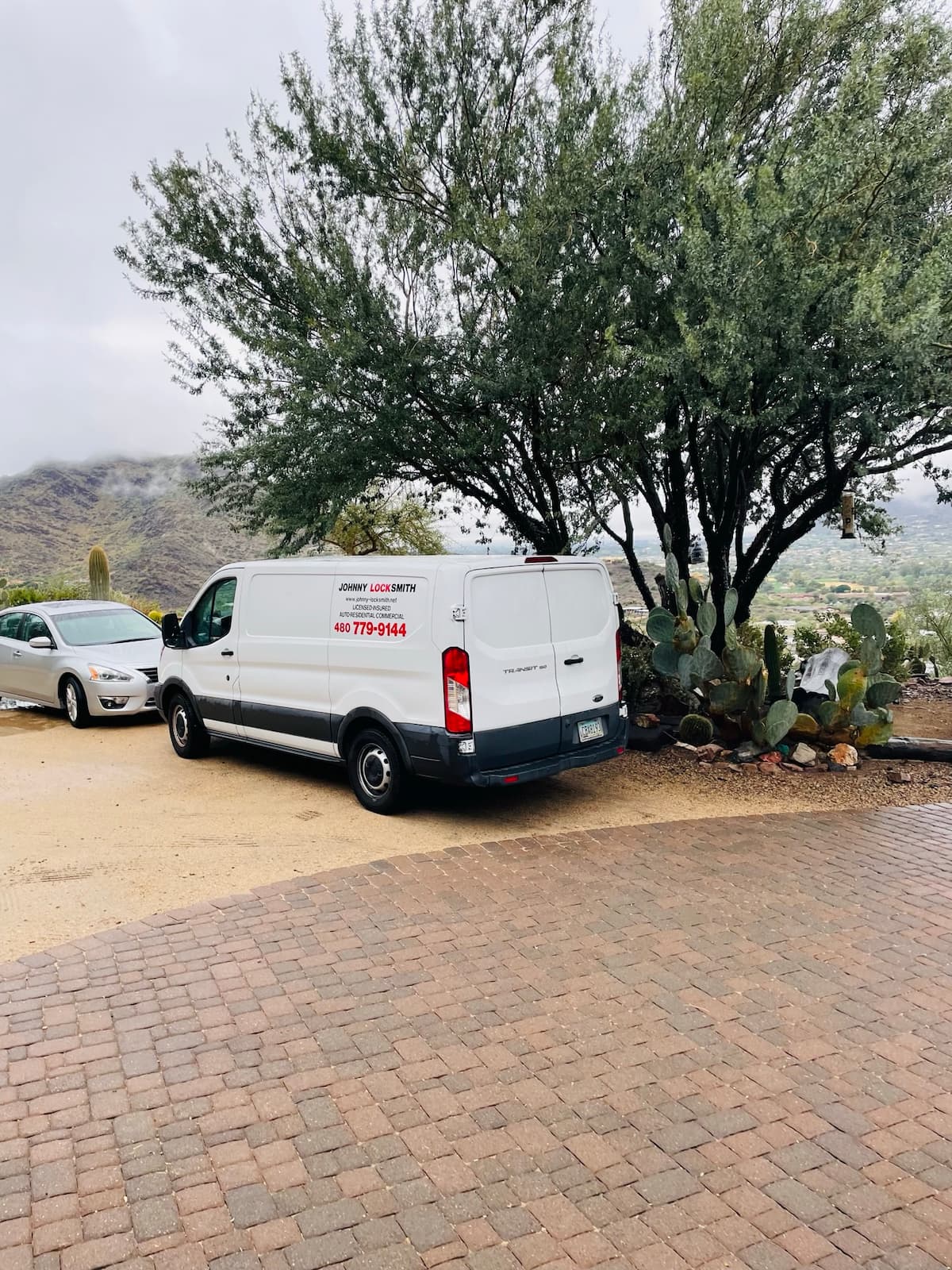 White van with Johnny Locksmith branding parked beside a large tree and cactus plants on a hill overlooking a valley.